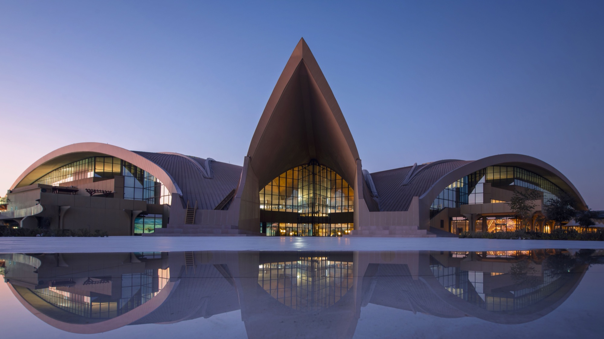 Sail-form landmark building with sweeping arched roofs mirrored in a reflecting pool at dusk